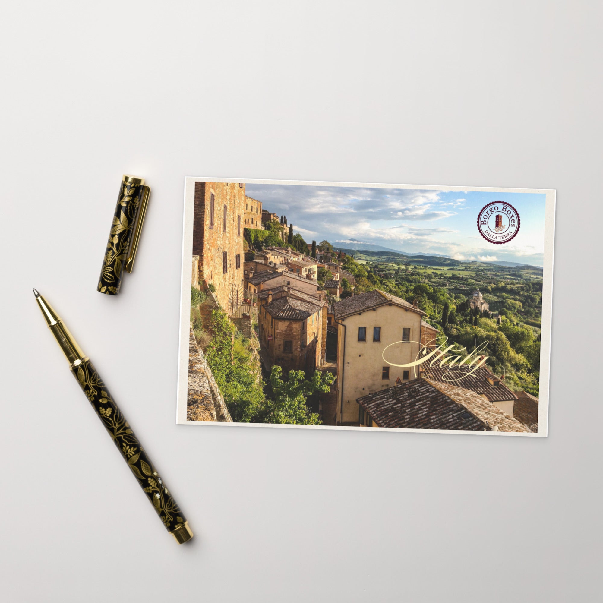 An ornate pen rests with the cap off next to a postcard from southern Italy to include in a gift basket. 