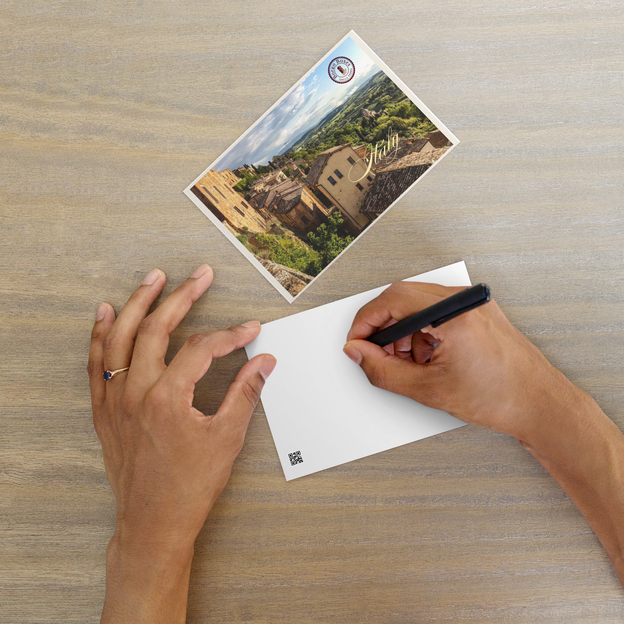 A hand is writing a note on a postcard from Italy to include in a gift basket. 